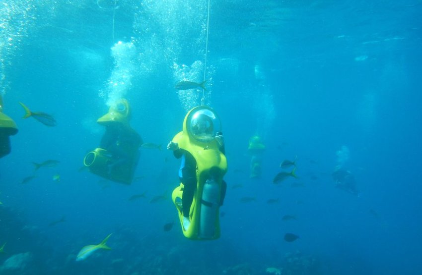 Personas viajando en scooters amarillos bajo el agua con peces por todas partes